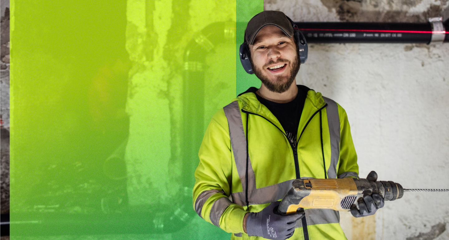 Smiling person in a high-visibility jacket, cap, ear protection, and gloves holding a power drill at a worksite