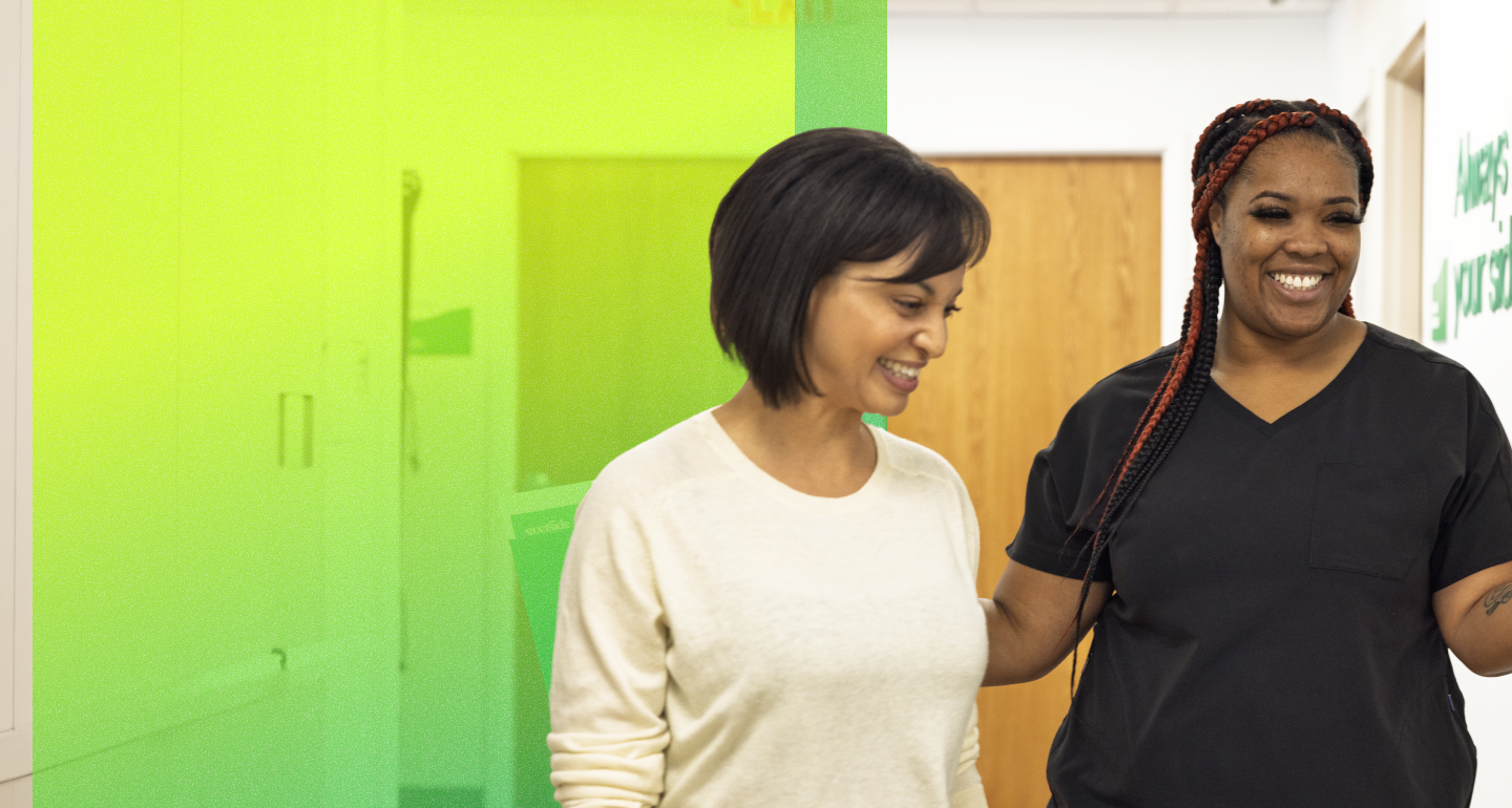 Two smiling people walk together down a bright hallway, one wearing dark scrubs and the other a light sweater