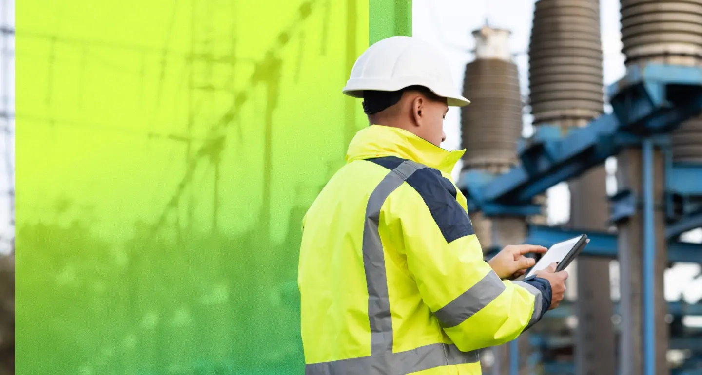 A person in a yellow high-visibility jacket and white hard hat holds a tablet at an industrial facility