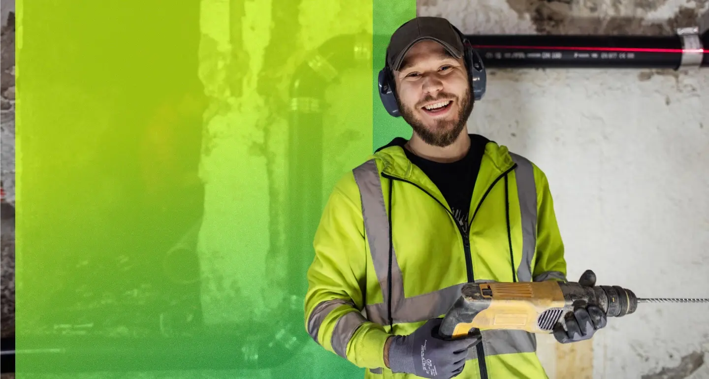 Smiling person in a high-visibility jacket, cap, ear protection, and gloves holding a power drill at a worksite