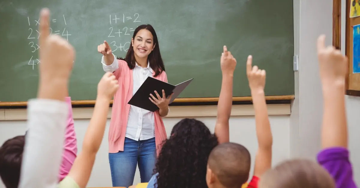 School teacher pointing at a group of students raising their hands.