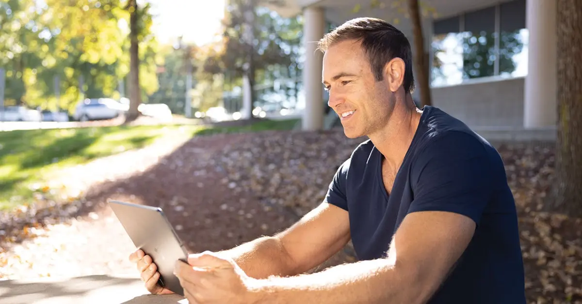 Smiling man sitting outside reading something on a tablet device.