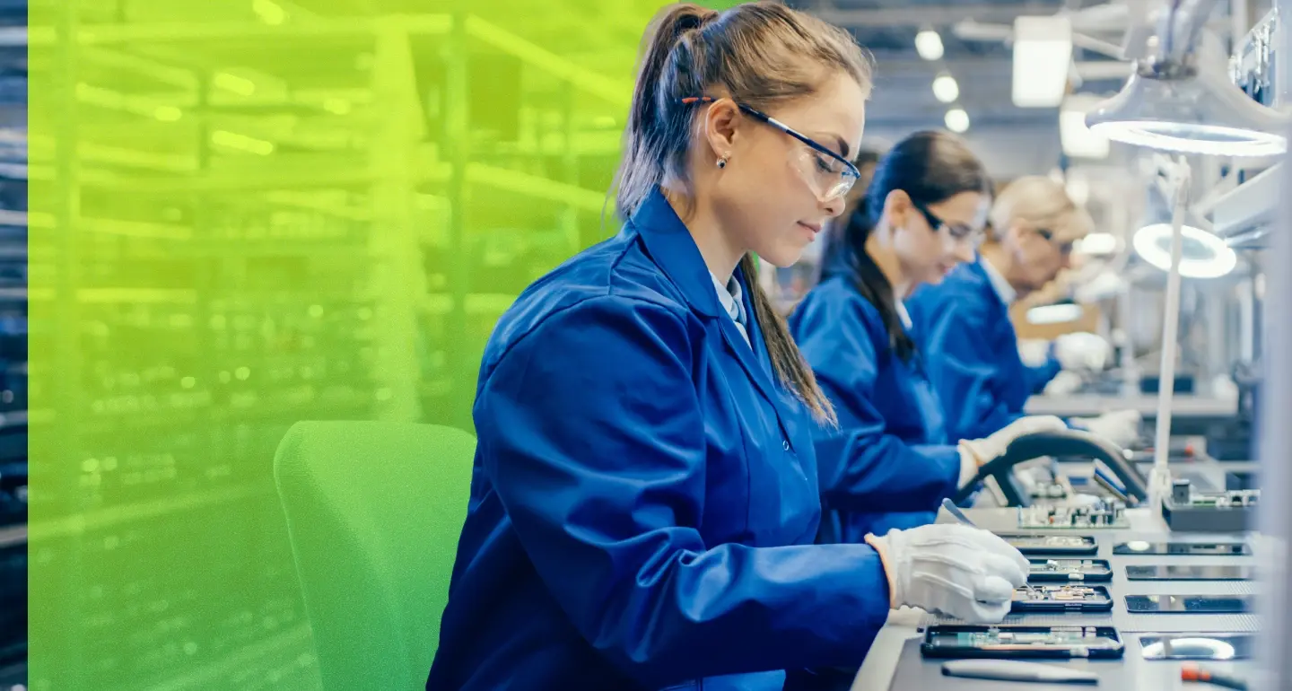 People in blue lab coats and safety glasses assemble circuit boards at a manufacturing workbench
